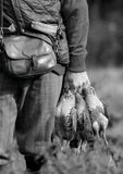 A black-and-white image from Countryside Greetings’ “10 Shooting notecards with envelopes by Charles Sainsbury-Plaice” shows a person in outdoor gear holding three game birds and a leather bag, reflecting country sports tradition.
