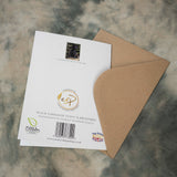 An A5 Black Labrador Dog Greeting Card (Puppy on Hay Bale) and a brown envelope from Countryside Greetings rest on a marbled surface, featuring eco-friendly symbols and photography by Charles Sainsbury-Plaice.