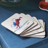 A stack of CountrysideGreetings Farm Animal Drinks Coasters by Alex Underdown rests on a blue surface; the top coaster shows a cow in red boots and blue scarf kicking up its legs. Made from sustainable cork for an eco-friendly touch.