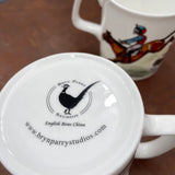 Close-up of the bottom of a CountrysideGreetings Bone China Horse Racing Mug (National Hunt Racing) showing the logo and website. Another mug with a National Hunt jockey illustration appears in the background, both on a brown surface.