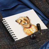 The Countryside Greetings Innocent by Bryn Parry cartoon dog-themed A6 lined notebook, featuring a Border Terrier in a red collar, is shown with a pen in the pocket of a blue satin-lined tweed coat.