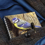 A Bird themed A6 lined notebook, Blue Tit by Dick Twinney from Countryside Greetings, and a pen are tucked into the pocket of a brown tweed coat with a blue lining.