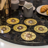 The Shotgun Cartridge Drinks Coaster Set by Charles Sainsbury-Plaice for Countryside Greetings features six round coasters designed like shell bases, displayed on a dark table with coffee cups and croissants—an ideal shooting gift.