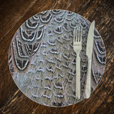 A metal fork and knife rest on a Countryside Greetings Decorative Placemat, Hen Pheasant Plumage by Charles Sainsbury-Plaice, featuring a feather-inspired design atop a wooden table.